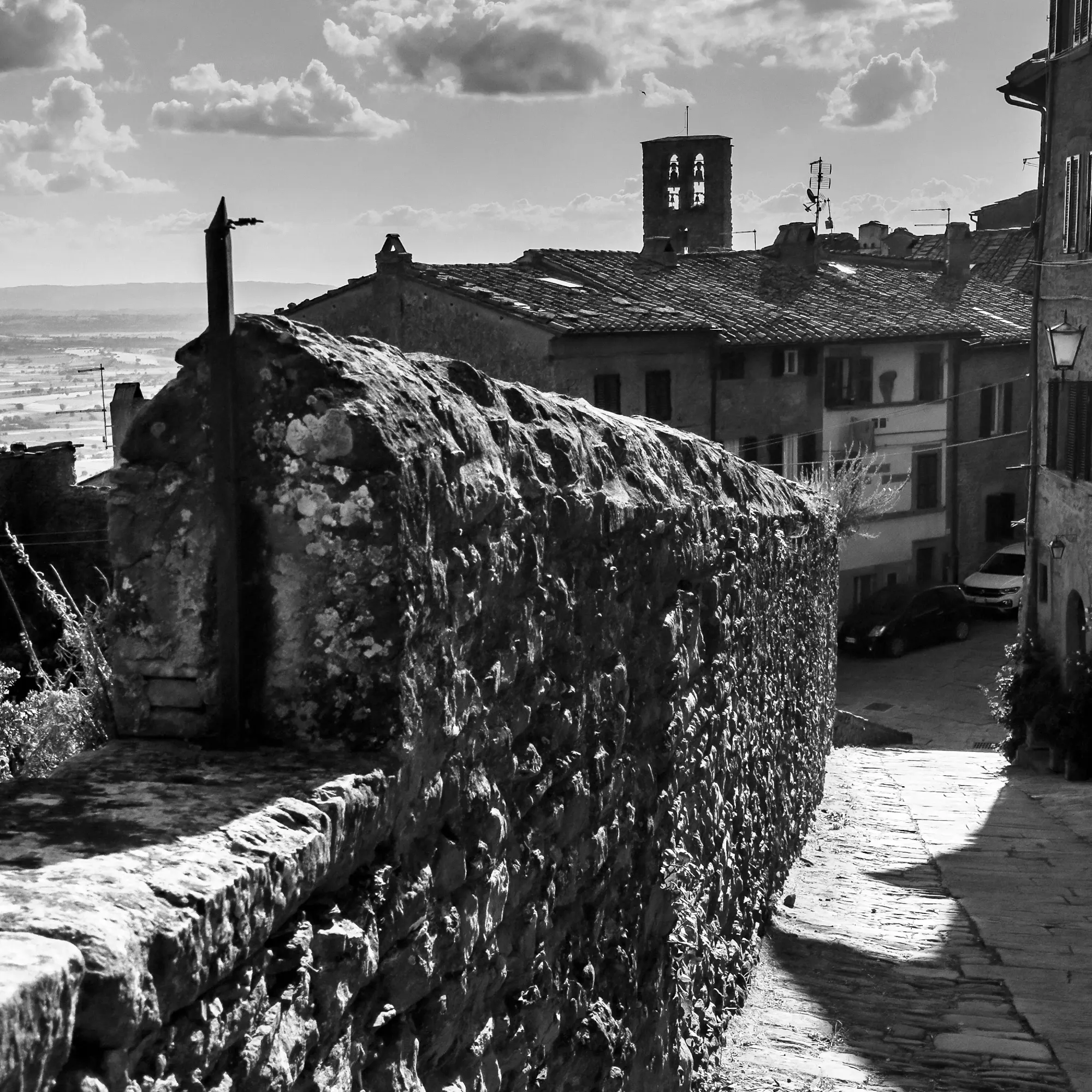 Monochromatic digital photograph of a stone wall and descending street in Cortona, Italy.